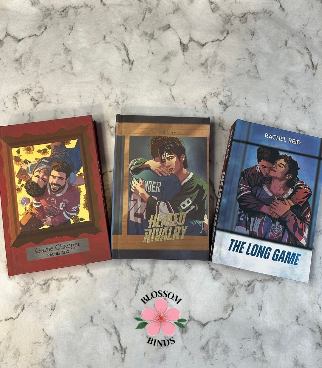 Three books titled 'Game Changer', 'Heated Rivalry', and 'The Long Game' on a marble surface with 'Blossom Binds' branding.