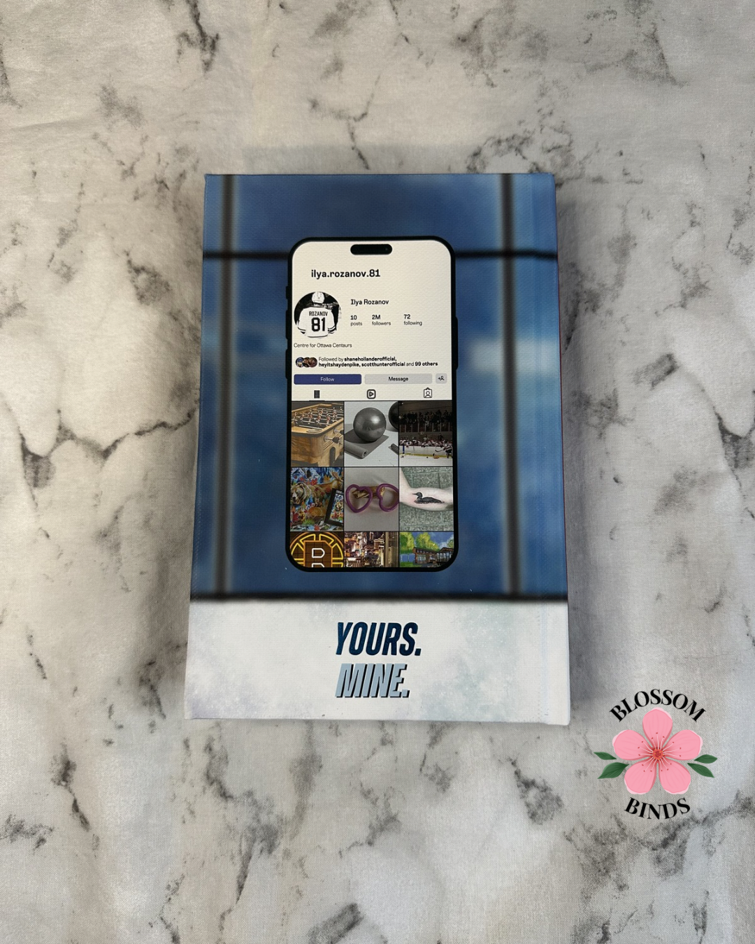 Book with social media app interface on marble surface, 'Blossom Binds' brand visible